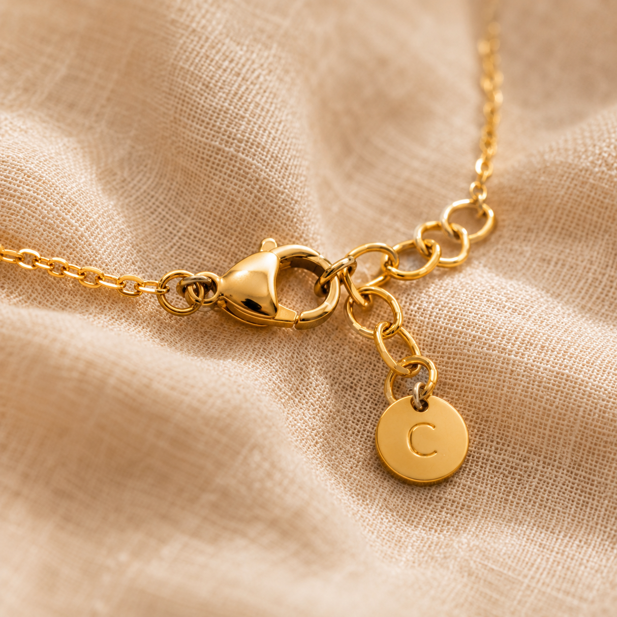Macro du fermoir ajustable doré d’un collier personnalisé avec médaillon gravé C sur fond lin beige premium.