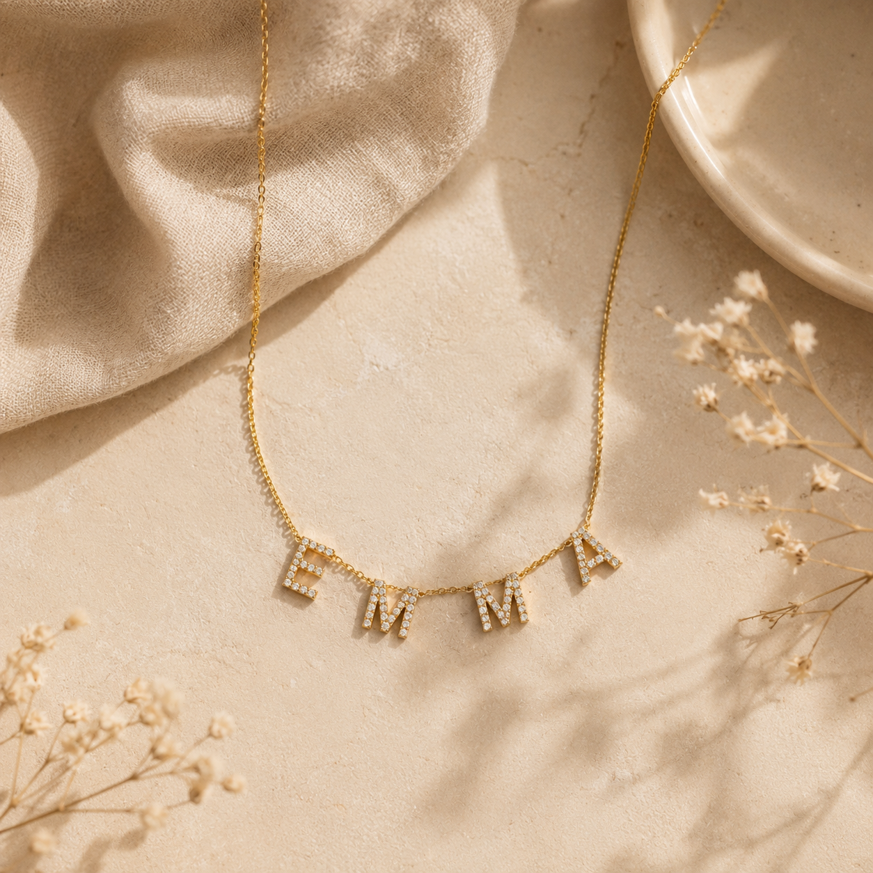 Collier personnalisé prénom EMMA en lettres dorées serties de strass posé sur un fond crème texturé avec lin beige et fleurs séchées.